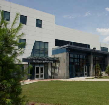 A modern, multi-story building with "Technology Building" above its entrance, surrounded by green grass and some trees under a partly cloudy sky.