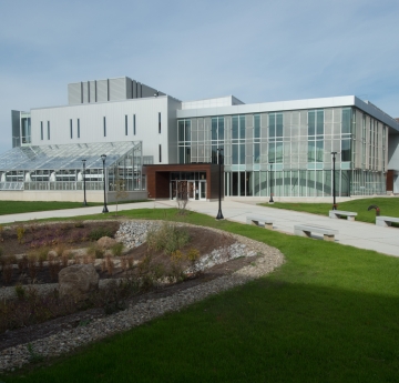 Science & Mathematics Complex -  A modern, large glass and concrete science building with an attached greenhouse is surrounded by green lawns, paved walkways, and a rock garden under a partly cloudy sky.
